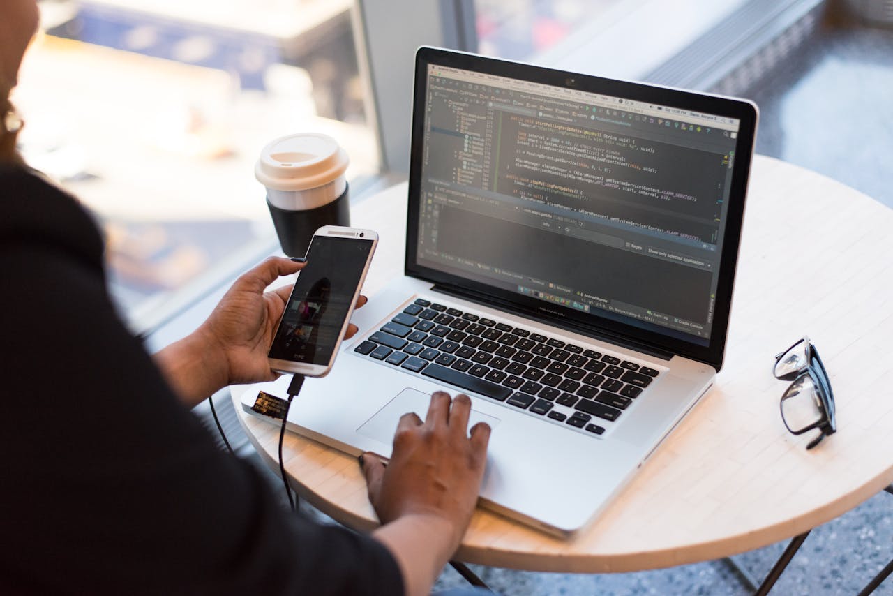 Home Developer working remotely, coding on a laptop with phone in hand, showcasing modern work culture.